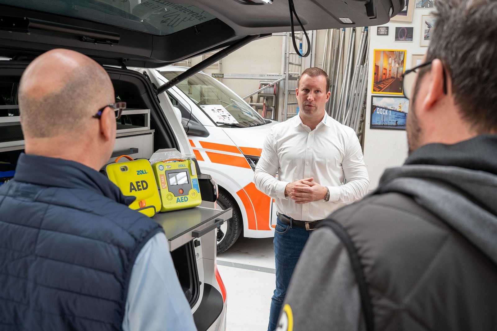 Ein ZOLL AED auf einer Ablage, dahinter ein Einsatzfahrzeug und zwei Personen im Gespräch